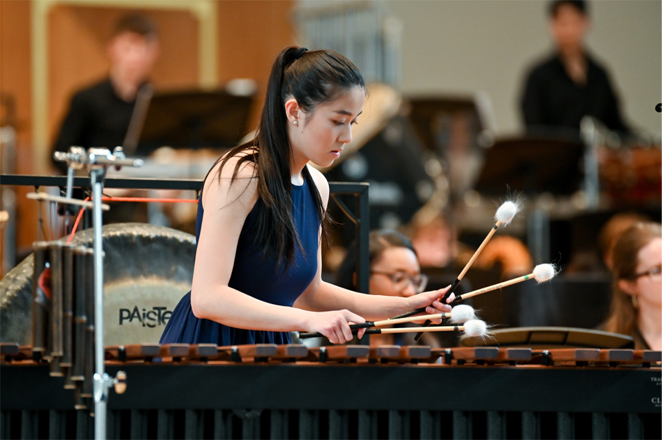 A women, wearing formal attire, performing on a marimba, in an orchestra performance.